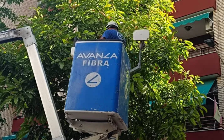 Operario con casco en el cesto de una grúa elevadora rotulada Avanza Fibra, junto a una farola y entre árboles, frente a un bloque de viviendas