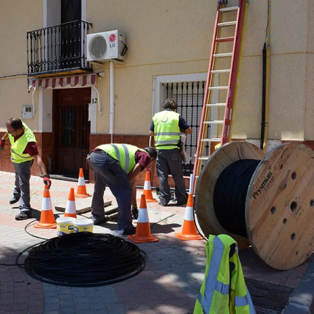 Tres operarios con chalecos reflectantes tienden cable de fibra óptica en una calle, junto a conos, una escalera y una bobina de cable