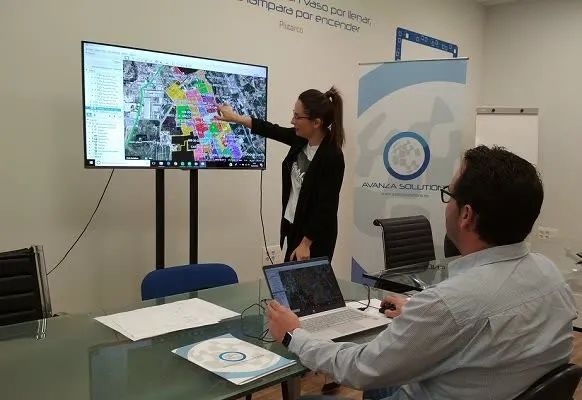 Dos técnicos en una sala de reuniones revisan en una pantalla y en un portátil el plano de una red sobre una vista aérea de la ciudad