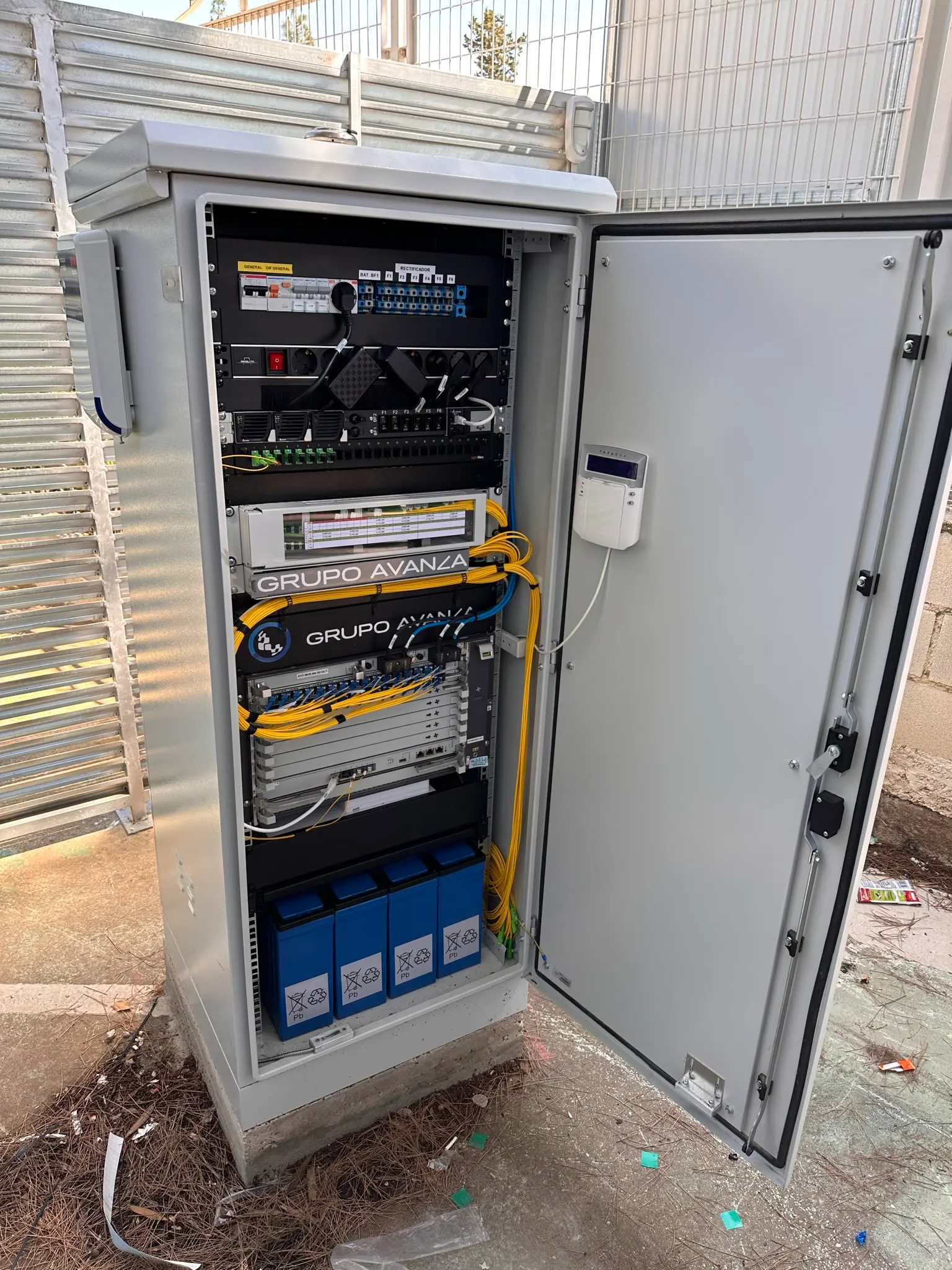 Armario exterior abierto con equipos de red, cableado de fibra amarillo y baterías, instalado sobre una base de hormigón